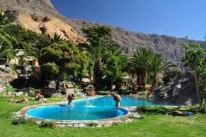 People at a large fountain or pool with many trees with buildings and steep cliffs.