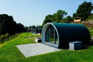 Modern glamping pods with curved roofs set on a green hillside surrounded by trees and landscaped grounds.