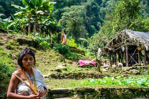 Woman wearing colorful traditional bead necklaces standing in a lush tropical village surrounded by banana trees, green vegetation, stone pathways, and rustic thatched-roof shelters.