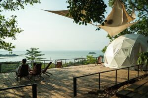Scenic glamping dome on a wooden deck overlooking calm water, surrounded by trees with guests relaxing outdoors.