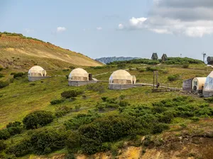 Scenic hillside glamping retreat featuring several cream-colored geodesic dome accommodations on raised platforms, surrounded by green vegetation, rolling hills, and distant mountain scenery.