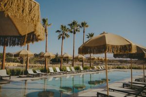 Luxury Tents in Huacachina Oasis: Desert Glamping & Adventures in Peru 5 Desert resort swimming pool surrounded by palm trees, thatched umbrellas, and comfortable lounge chairs beneath a clear blue sky.