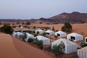 Luxury Tents in Huacachina Oasis: Desert Glamping & Adventures in Peru 2 Luxury desert glamping camp featuring white safari-style tents surrounded by golden sand dunes, scattered trees, and distant mountains.