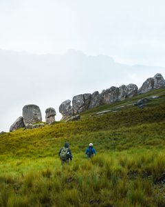 Two hikers carrying backpacks walk through tall green mountain grass toward unusual rock formations on a rugged hillside beneath a cloudy sky, illustrating scenic guided mountain trekking adventures.