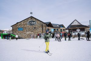 La Parva Mountain Retreat: A High-Altitude Escape in the Andes 1 Skiers enjoying a snowy mountain resort plaza with a stone-built restaurant, ski equipment racks, alpine buildings, and spectacular snow-covered mountain peaks. A skier wearing a green jacket and bright yellow pants glides across the snow beneath a clear blue sky.