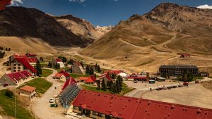 Glamping In Cajón Del Maipo: Andes Luxury Camping Near Santiago, Chile 2 Panoramic view of a mountain resort village with red-roofed lodges, hotels, winding roads, and rugged Andes Mountains beneath a clear blue sky.