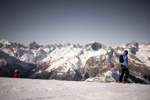 La Parva Mountain Retreat: A High-Altitude Escape in the Andes 5 Skier wearing a blue jacket and carrying ski poles standing on a snowy mountain slope, with another skier in red and spectacular snow-covered alpine peaks in the background.