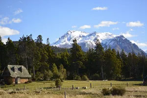 Rustic wooden mountain cabin overlooking a grassy meadow surrounded by dense evergreen forests, with towering snow-covered Andean mountain peaks rising beneath a bright blue sky.