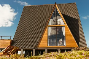 Modern A-frame wooden mountain cabin featuring a steep dark roof, large triangular floor-to-ceiling windows, white curtains, and an elevated wooden deck, surrounded by low vegetation beneath a bright blue sky.