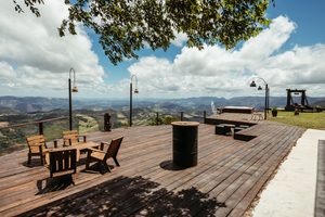 Spacious wooden observation deck featuring rustic outdoor tables, chairs, decorative lanterns, and panoramic views of rolling green hills and distant mountains beneath a partly cloudy sky.