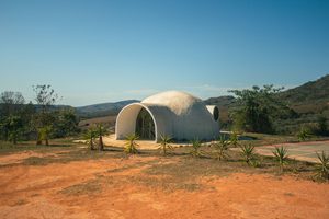 Unique white dome-shaped eco-building surrounded by rolling countryside hills, small tropical plants, reddish earth, and clear blue skies in a peaceful natural landscape.