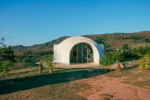 White dome-shaped retreat with a decorative arched entrance, surrounded by green vegetation, palm-like plants, and rolling mountains beneath a clear blue sky.