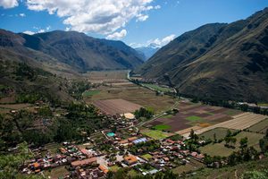 Panoramic view of Peru's Sacred Valley featuring towering Andes Mountains, green agricultural fields, traditional village buildings, and a winding river beneath a bright blue sky.