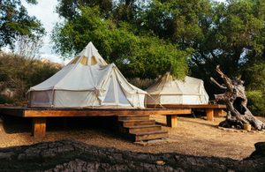 Two spacious canvas glamping tents on elevated wooden platforms with steps, surrounded by mature trees and natural woodland scenery, illustrating a peaceful countryside camping retreat.