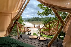 Guararema Riverside Glamping, Brazil: A Peaceful Escape by the Water 5 Riverside glamping tent with two wicker chairs on a wooden deck overlooking a peaceful river, lush green forests, and distant hills beneath a cloudy sky.