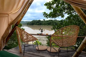 Riverfront Dome, Guapimirim – Tranquil Jungle Retreat By The River 3 Cozy riverside glamping deck featuring two wicker chairs, a small coffee setup, and beautiful views of a peaceful river surrounded by lush tropical forest in Guapimirim, Brazil.