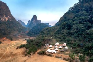 Aerial view of a remote glamping retreat featuring white safari tents, small cabins, and outdoor gathering areas surrounded by towering forest-covered mountains and dramatic rocky peaks.