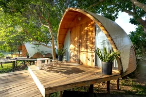 Riverfront Dome, Guapimirim – Tranquil Jungle Retreat By The River 2 Text: Modern arched wooden glamping dome surrounded by lush tropical trees in Guapimirim, Brazil, featuring large glass windows, a private wooden deck, outdoor seating, and peaceful jungle scenery.