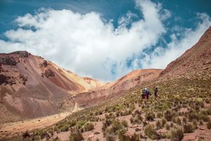 La Parva Mountain Retreat: A High-Altitude Escape in the Andes 6 Three hikers carrying backpacks along a rugged mountain trail surrounded by reddish-brown Andean slopes, sparse alpine grasses, and dramatic clouds against a blue sky.