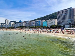 ✨ Rio De Janeiro Glamping Hub: Luxury Nature Stays in Brazil’s Most Vibrant City 4 Swimmers enjoying the Atlantic Ocean at Copacabana Beach in Rio de Janeiro, Brazil, with colorful beach umbrellas, beachfront hotels, and green mountains in the background.