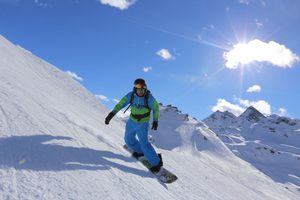 La Parva Mountain Retreat: A High-Altitude Escape in the Andes 4 Snowboarder wearing a blue and green winter outfit descending a steep, snow-covered mountain slope, with rugged alpine peaks and bright sunshine in the background.