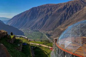 Glamping dome made of large windows on a hill overlooking a valley with a tall mountain in the background.