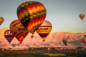 Several hot air balloons flying over fields with mountains in the background,