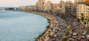 🌊 Alexandria – Mediterranean Charm, Libraries & Coastal Fortresses 1 Buildings and cars on a highway near the ocean with people.
