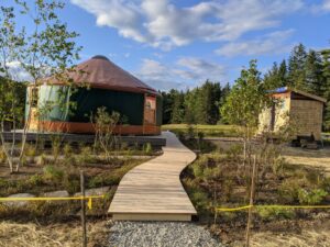 Brown glamping yurt on a raised platform, with a wooden pathway leading to it, surrounded by sandy fields and scattered trees