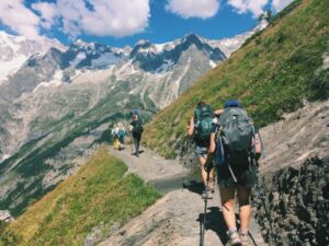 Group of hikers climbing a steep mountain trail in the Alps, carrying backpacks and trekking poles, surrounded by rugged peaks and scenic landscapes.