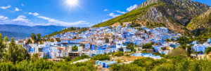 Chefchaouen, Morocco – The Enchanting Blue City 1 Panoramic view of Chefchaouen, Morocco, featuring blue-and-white buildings nestled beneath the rugged Rif Mountains on a bright sunny day.