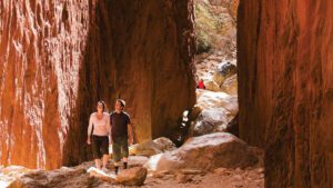 Two people walking through a narrow canyon passage with steep rocky walls on either side.