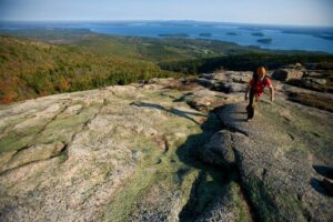 Person with a backpack hiking on a flat rock surface atop a hill, overlooking a forest and lake in the background.