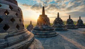 🌄 Explore Borobudur, Indonesia: Majestic Temples, Sunrise Views & Cultural Adventures 3 Several acorn-shaped stupas and a temple at Borobudur, Indonesia, surrounded by woods at sunrise.