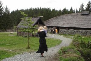 Viking Longhouses: Homes Of The NorseViking Longhouses Were The Central 3 Reconstructed Viking longhouse with three people outside, surrounded by grass and forest, demonstrating Viking-era architecture and lifestyle.