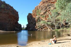 Person sitting on a sandy beach while others swim in a lake, surrounded by steep canyon walls and natural scenery.