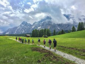 Thirteen hikers with backpacks walking on a stone path, with green grass and distant mountains.