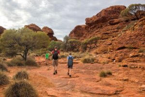 Three hikers, two wearing backpacks, walking among trees and large rock formations in a natural outdoor setting.