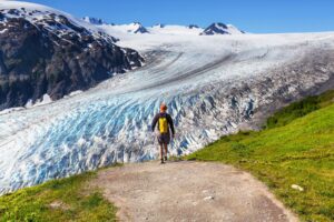 Person with a backpack walking along a trail toward a glacier surrounded by rugged terrain.