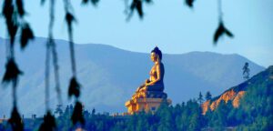 Large statue of a seated figure in a forested area with mountains in Bhutan.