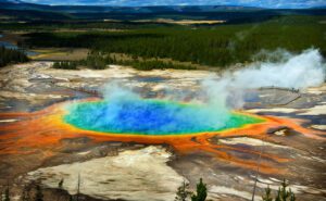 Large blue water geyser with an orange ring, set within a pine tree forest.
