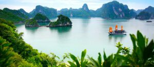 Boat floating on a calm lake surrounded by rainforest and tall, rounded rocky mountains.
