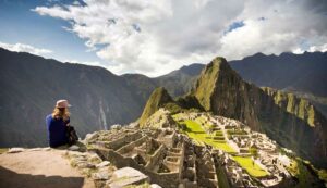 Person sitting on a ledge overlooking Machu Picchu, Peru, with terraces and temples surrounded by misty Andes mountains, perfect for a holiday or winter adventure.