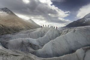 Several hikers walking across a distant glacier in the mountains, surrounded by snow, ice, and alpine peaks.