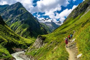 Hikers with backpacks walking up a mountain trail, with a creek flowing below and towering alpine peaks in the distance.