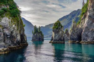 Scenic lake view – towering rock formations rise from the water with trees on top, mountains looming in the distance on a cloudy day.