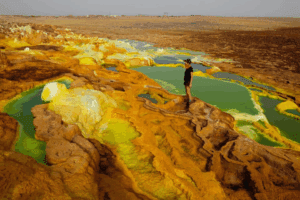 Danakil Depression – Ethiopia’s Otherworldly Landscapes 1 Person standing above colorful mineral pools and salt formations in Ethiopia’s Danakil Depression, one of the hottest places on Earth.”
