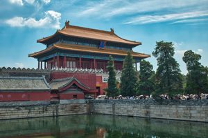 Historic Chinese palace building with golden tiled rooftops, red walls, decorative architectural details, and visitors walking beside a reflecting moat at the Forbidden City in Beijing, China.