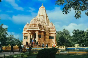 Ornate Hindu temple in India featuring a tall carved stone spire, detailed architectural decorations, visitors at the entrance, and landscaped gardens beneath a bright blue sky.
