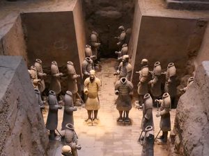 Ancient terracotta warrior figures arranged inside an earthen excavation chamber, with several headless clay soldiers surrounding a standing armored warrior.